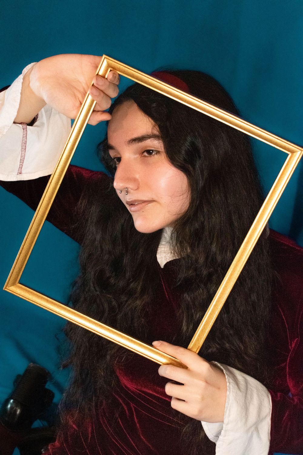 Portrait against teal background framed by gold picture frame with long dark hair and velvet burgundy top.
