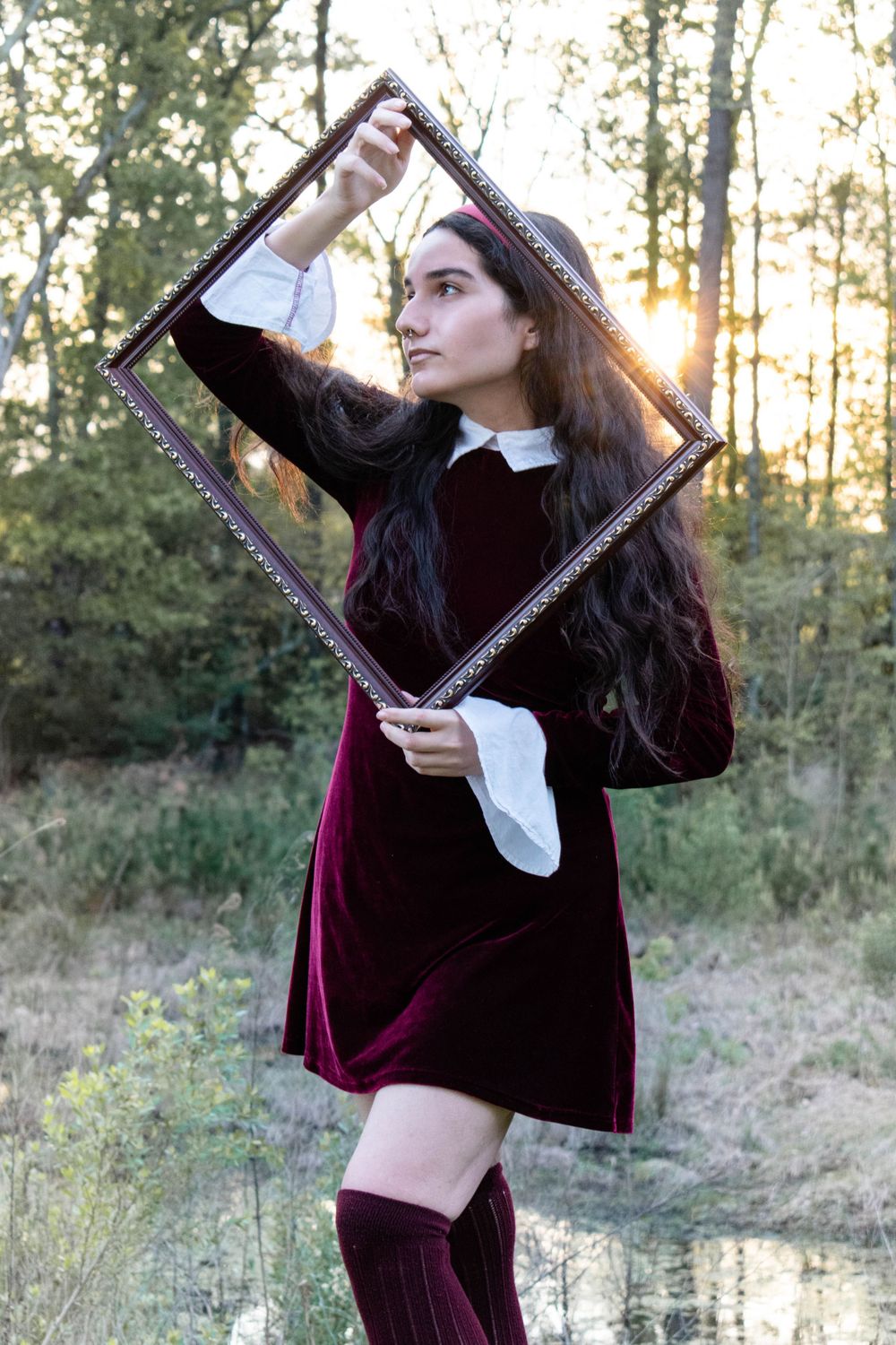 Figure in velvet dress holds ornate frame outdoors at sunset.