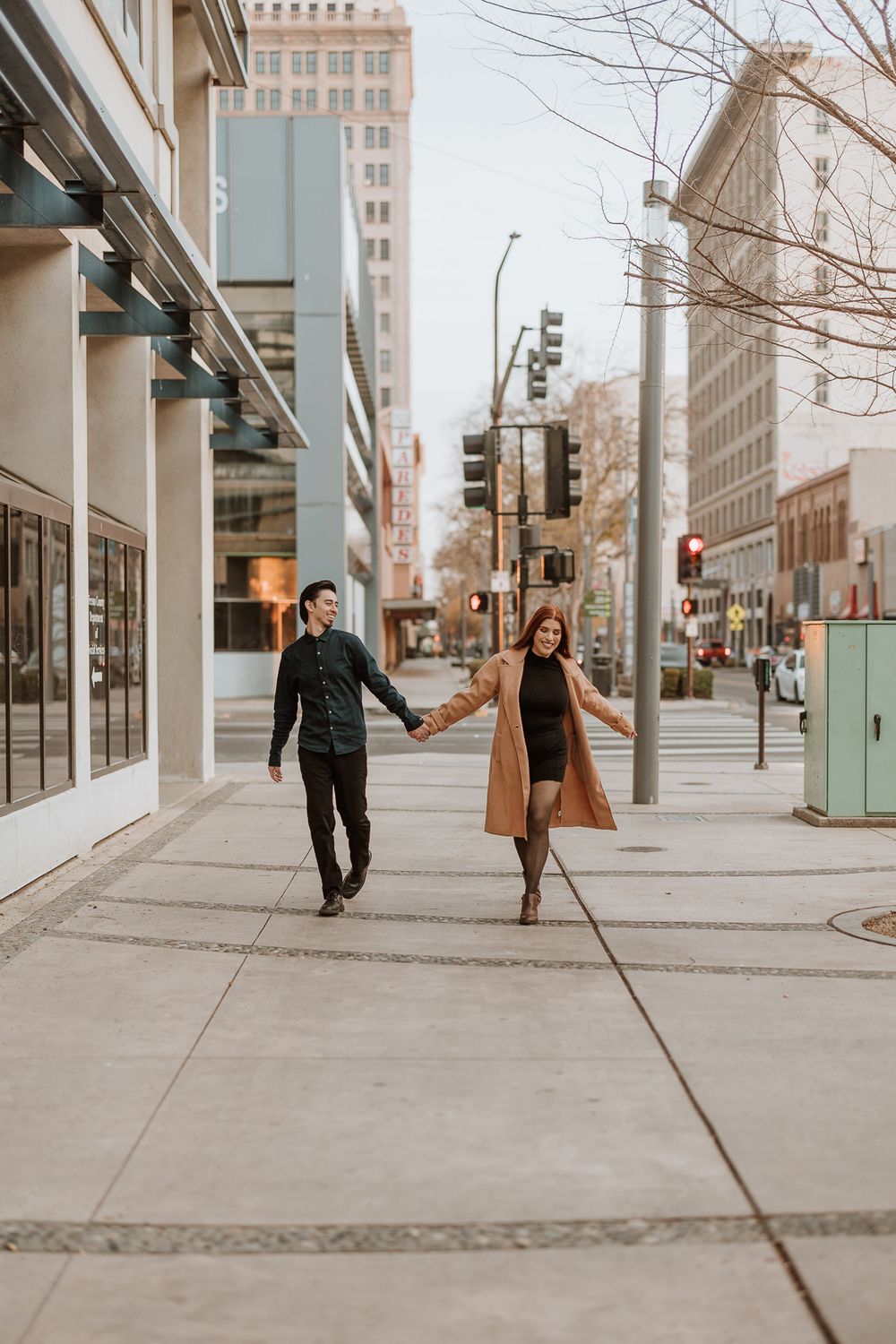 Couple walking hand in hand down city sidewalk with urban buildings in background.