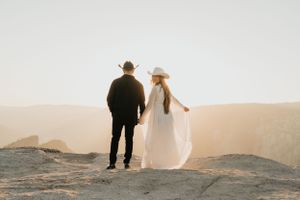 Couple in black and white attire standing on mountain edge at sunset in romantic desert landscape.