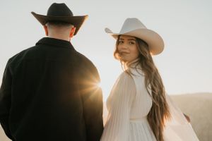 Couple in western attire with cowboy hats pose against glowing sunset in mountains.