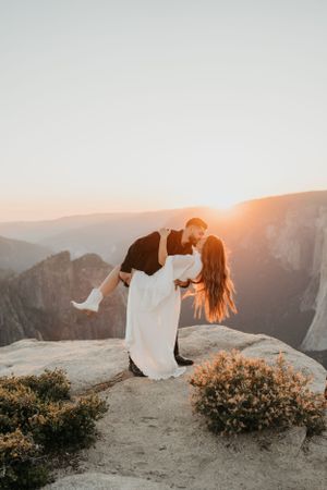 Romantic couple sharing intimate moment on cliff edge with stunning mountain sunset backdrop.