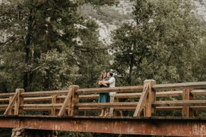 A couple embraces on a rustic wooden bridge surrounded by lush green trees.