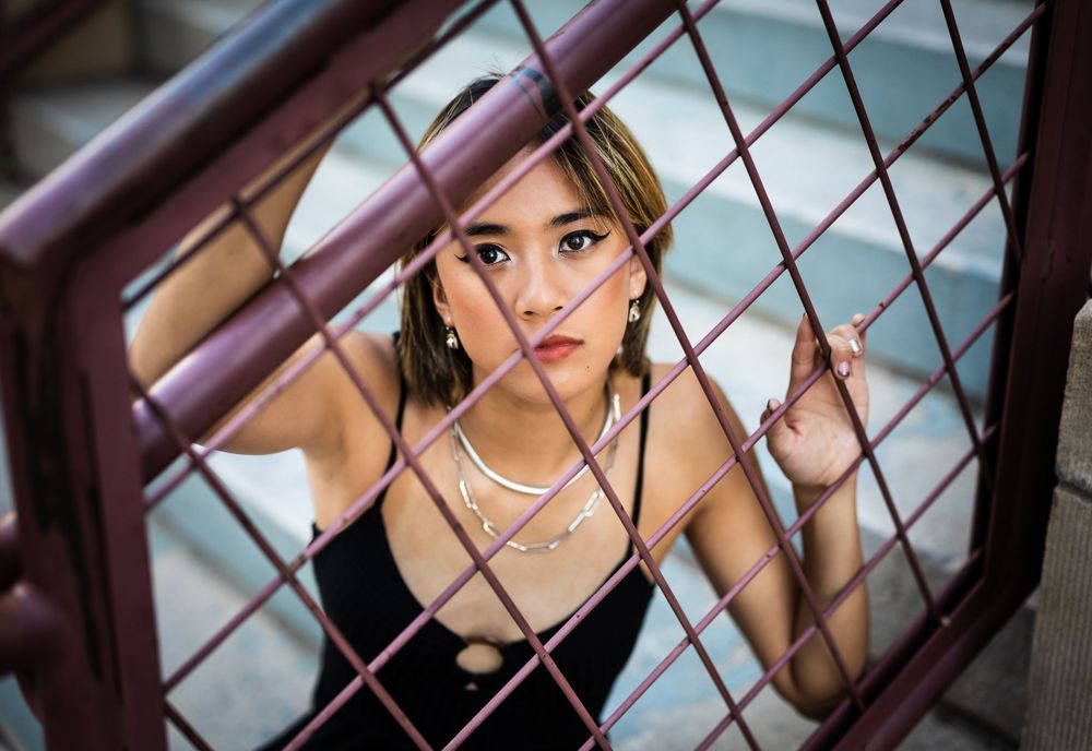 Artistic portrait through metal fence showing jewelry and dramatic lighting.