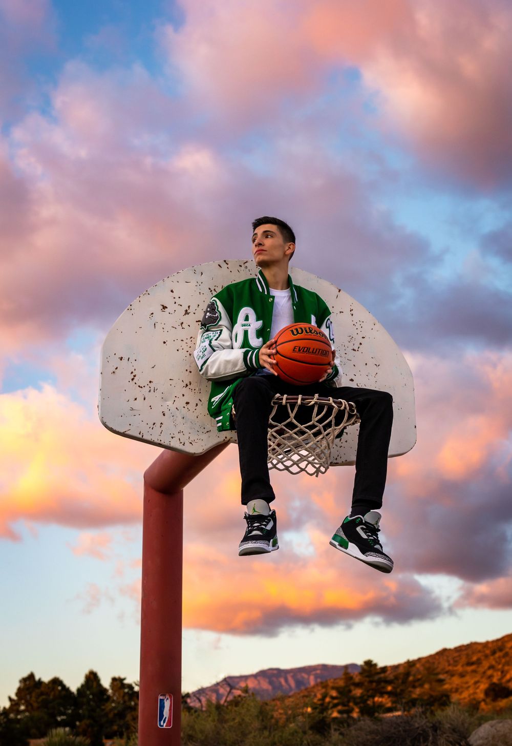 Basketball player sits atop hoop backboard against dramatic sunset sky.