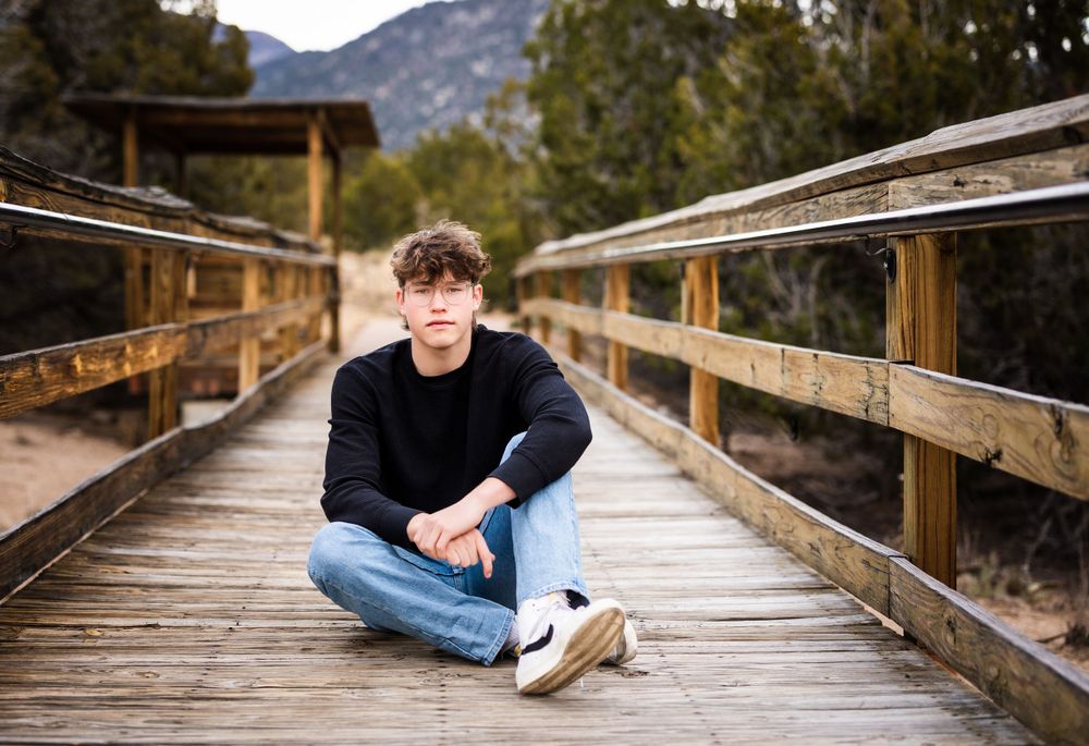 Casual portrait of person sitting cross-legged on wooden bridge with mountains behind.