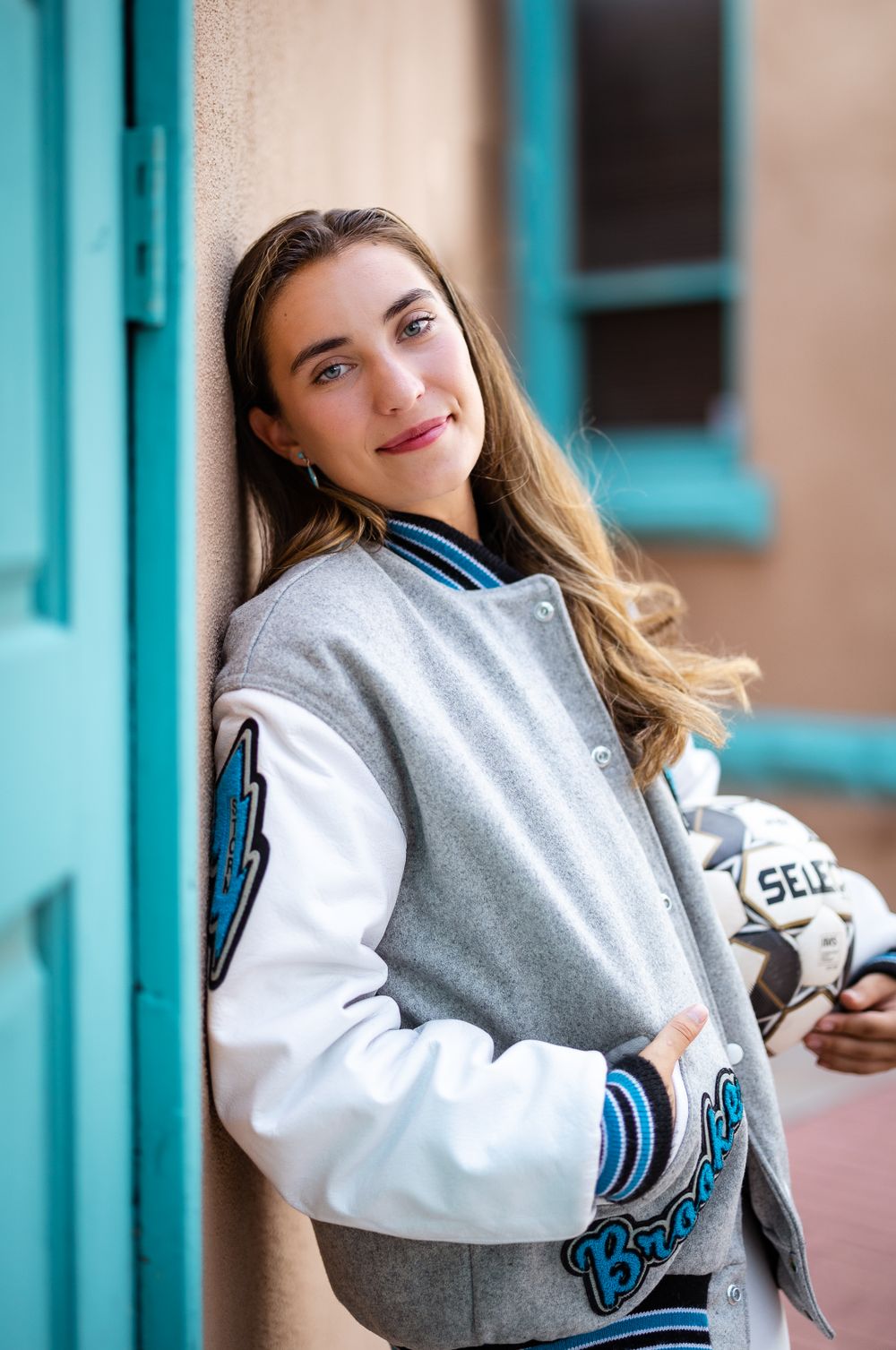 Casual portrait of person in grey and white varsity jacket leaning against turquoise wall.