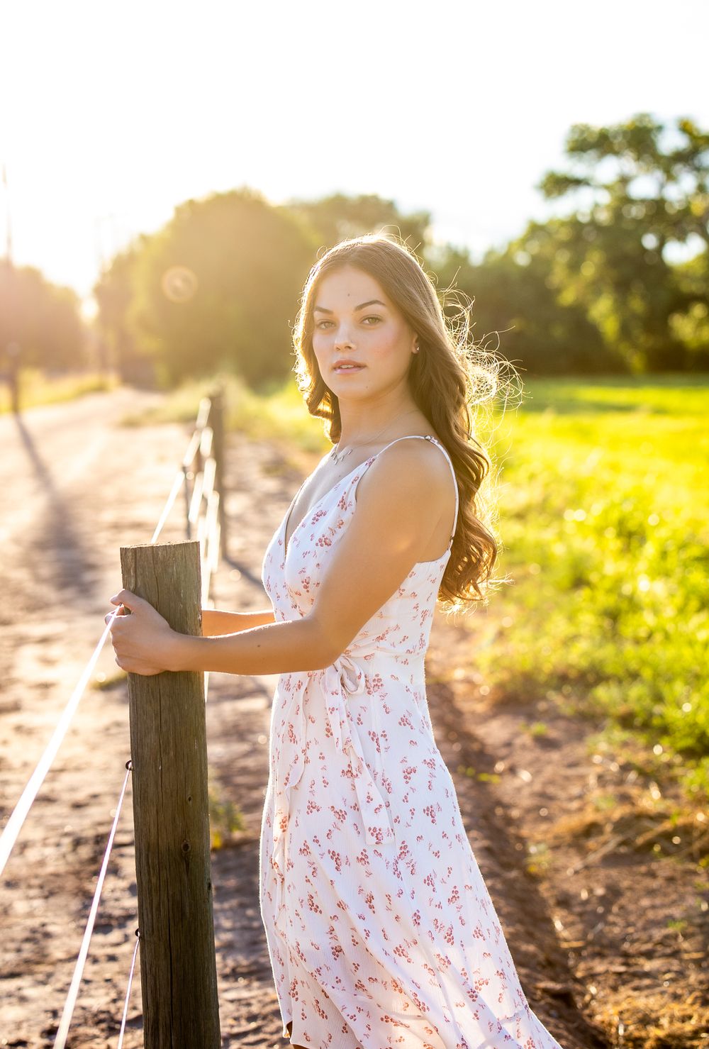 A backlit portrait of a woman in a white dress standing by a wooden fence post at golden hour.