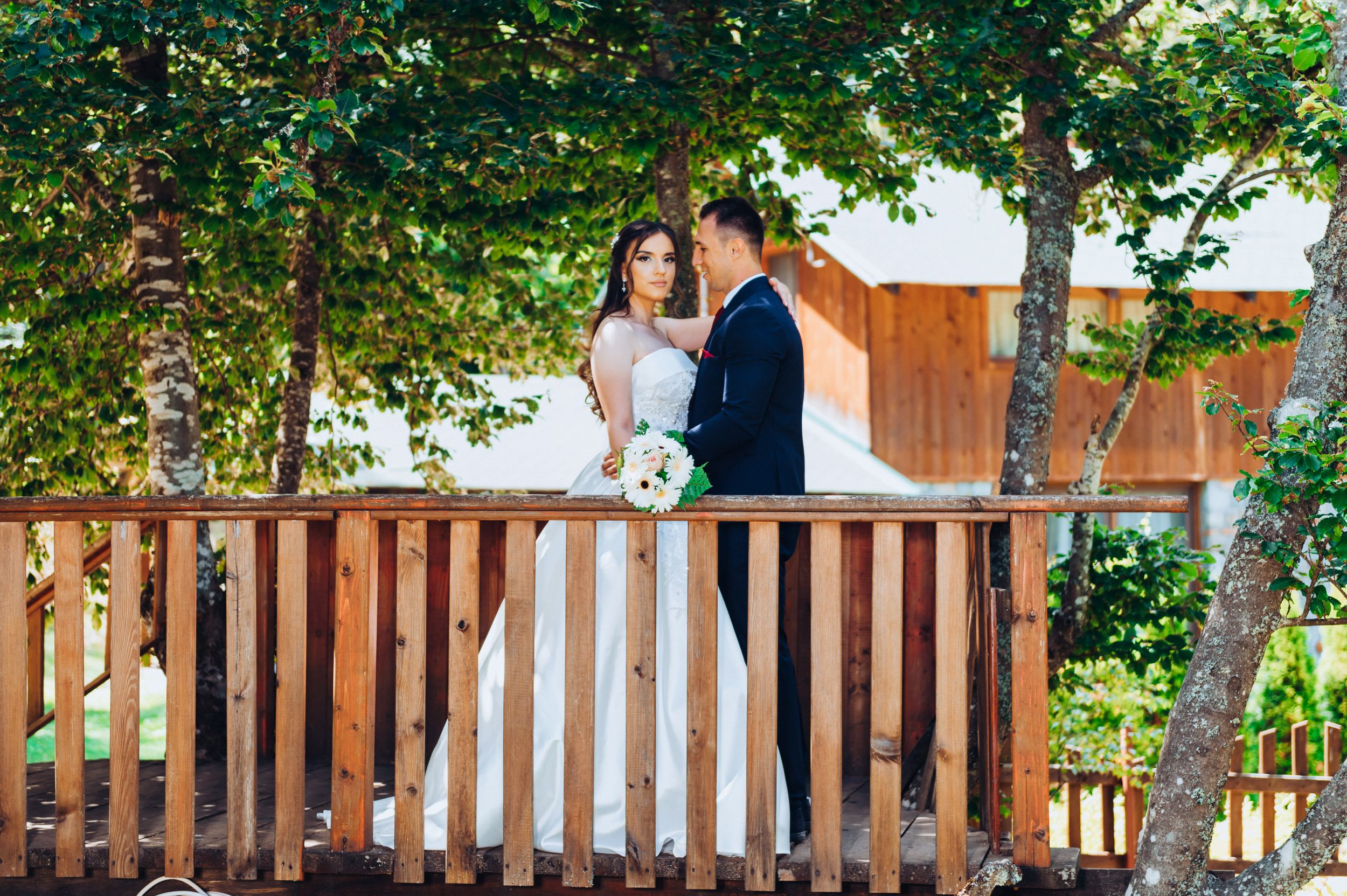 Wedding couple stands together on wooden balcony with lush greenery in background.