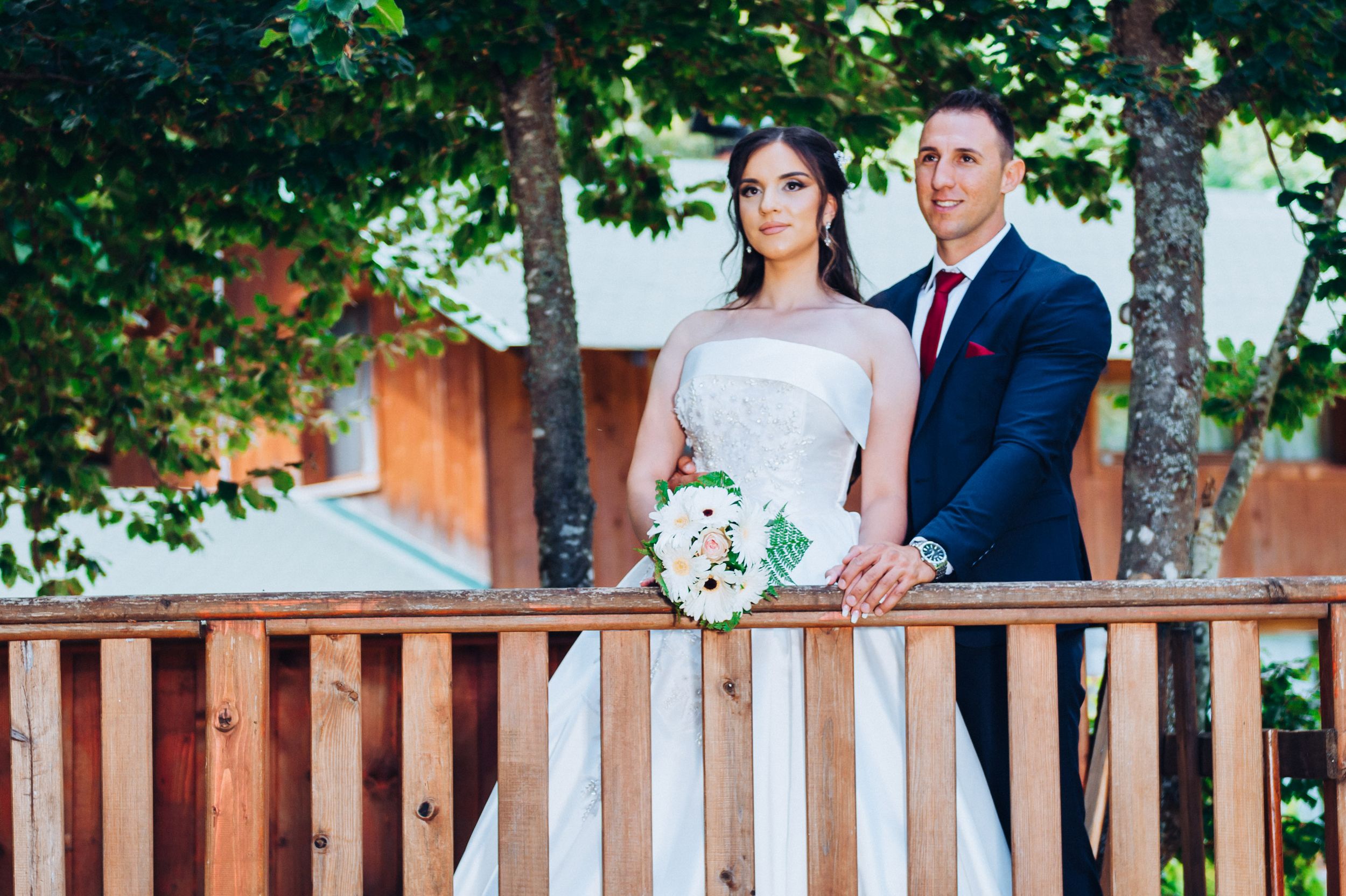 Newlywed couple poses on rustic wooden deck surrounded by green trees during outdoor wedding photoshoot.