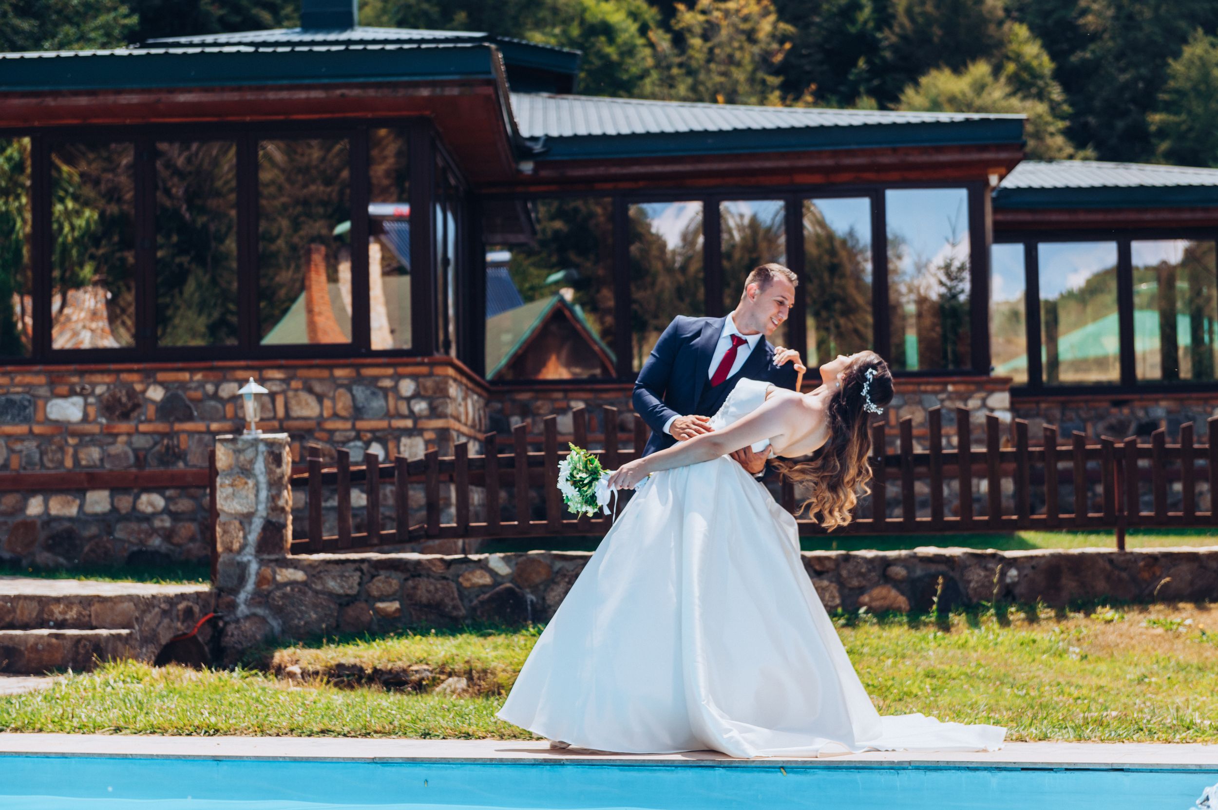 Romantic wedding couple embraces by pool with rustic pavilion in background on sunny day.
