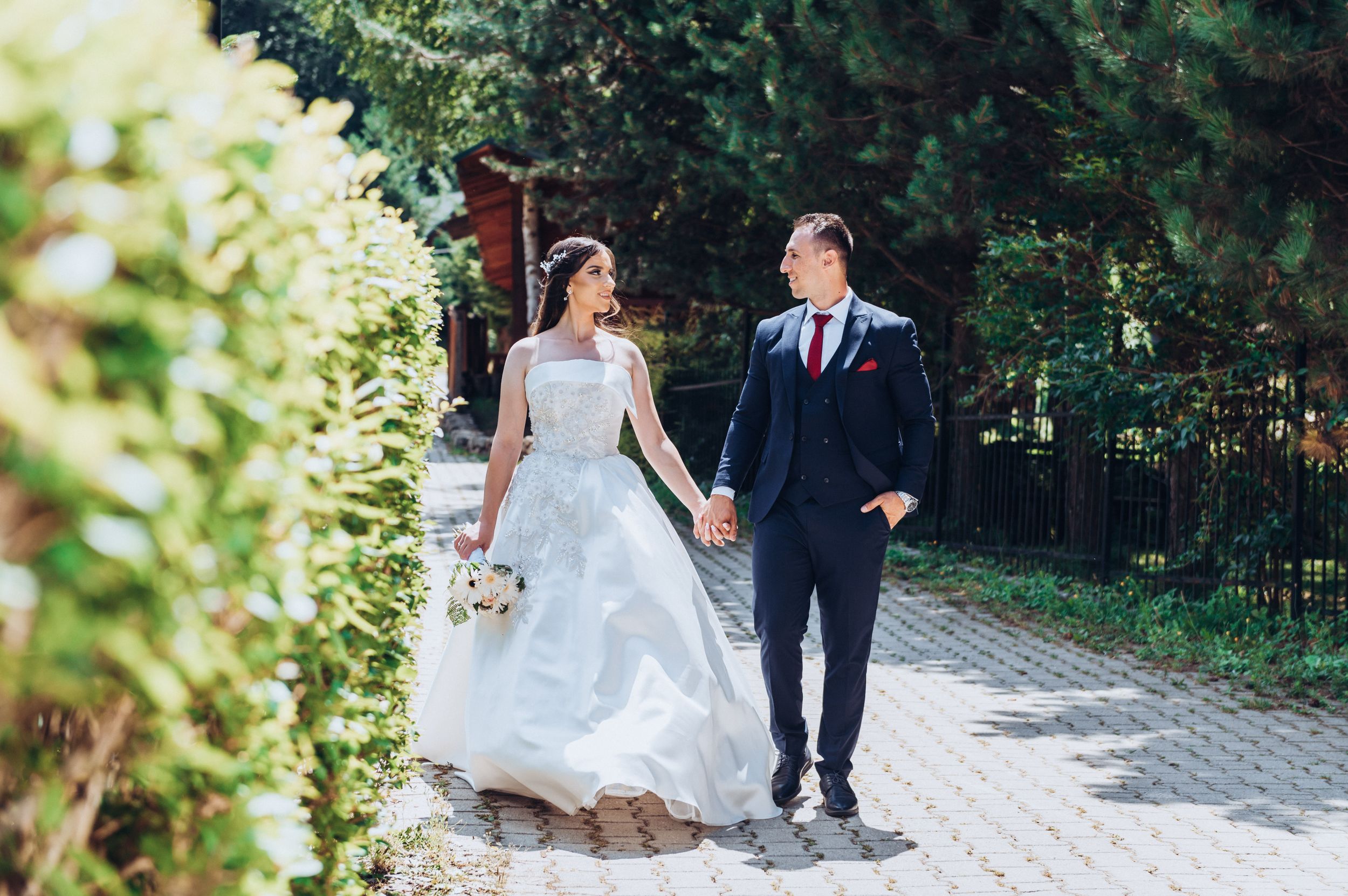 Bride in flowing white gown and groom in navy suit walk hand-in-hand along garden path.