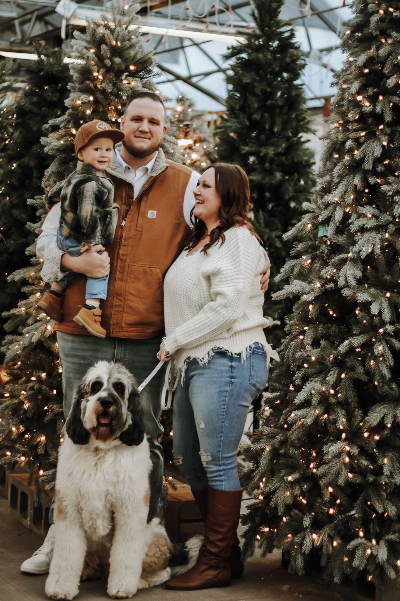 A family poses with their large fluffy dog among decorated Christmas trees and twinkling lights.