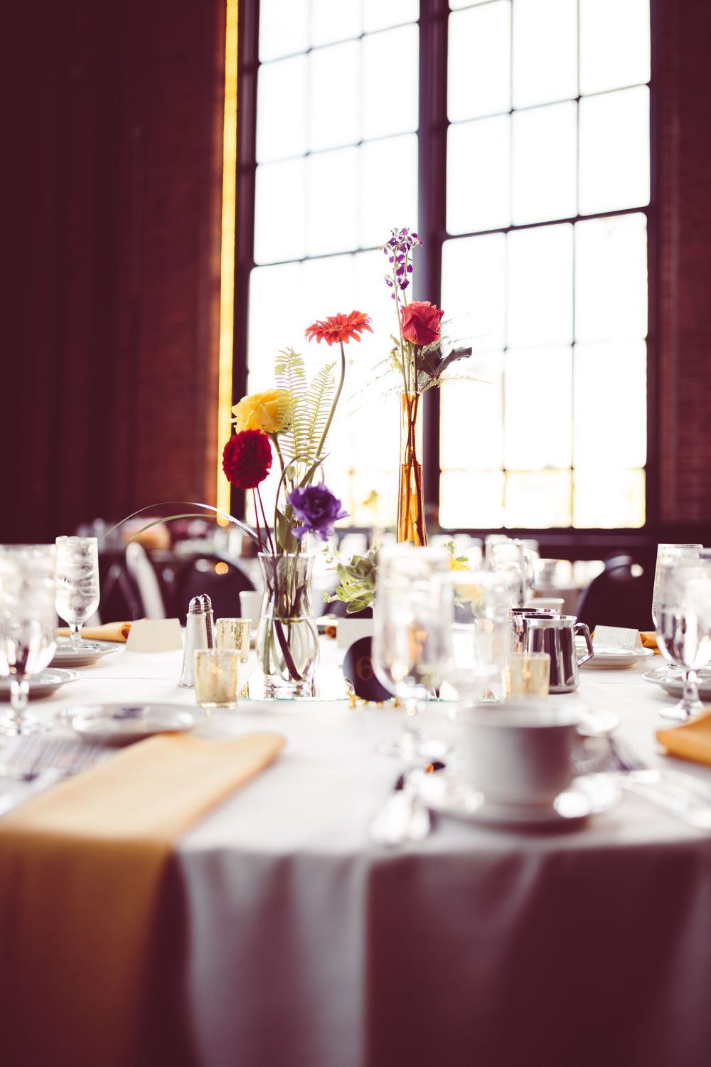 Elegant wedding reception tables set with white linens and floral arrangements near large windows.