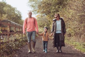 A young family are holding hands in a line, walking towards the camera, down a track. There is a steam train on the left side of the camera.