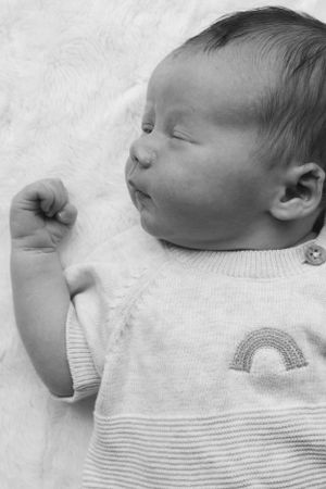 A black and white image of a newborn baby asleep. He has his arm up to the side by his face.