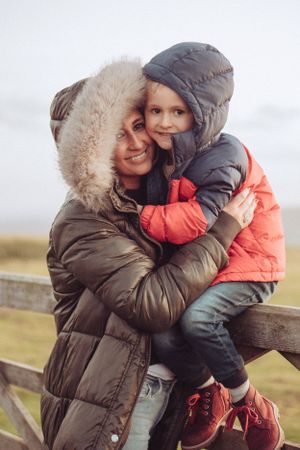 Mum hugging son who is sat on a gate. Both wearing winter coats with hoods up