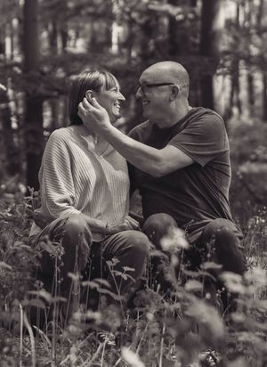 A black and white image of a married couple. They are sat on a log in a woods. He is tucking the hair behind her ear, whilst they look.