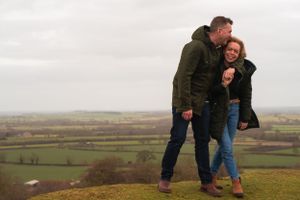 A married couple are having a dance and a cuddle on the hills with stunning views behind them. He is looking down at her and both laughing