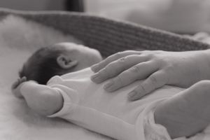 A black and white image of a newborn baby in a baby basket. Parents hand is resting on their tummy to settle them.