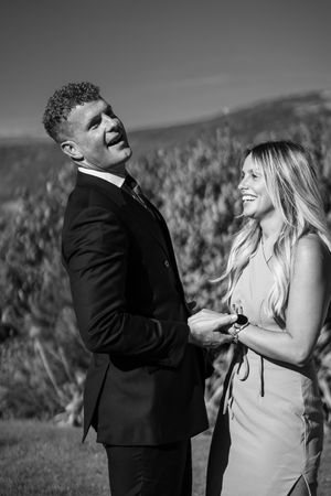 A black and white image of a young couple having a laugh. They are holding hands, she is looking at him and smiling, he is leaning back