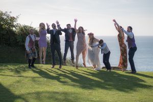 A wedding party are stood in front of the ocean and are all jumping. They are all mid air.