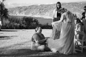 A bride has her back to the camera, one of her bridesmaids is sat on the floor altering the bottom of the brides dress. Black and white