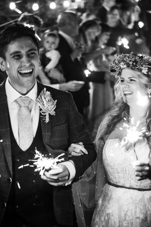 A black and white image of a newly wed couple. They are laughing, she is looking at him and they are holding sparklers!