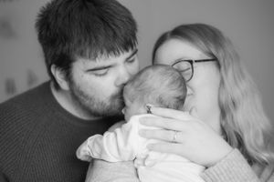 A black and white photo of new parents cuddling their newborn baby girl. Dad is giving the baby a kiss on the head.