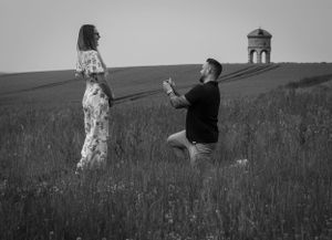 A black and white image of a man proposing! He is down on one knee and she is laughing! In an open field with a old windmill.