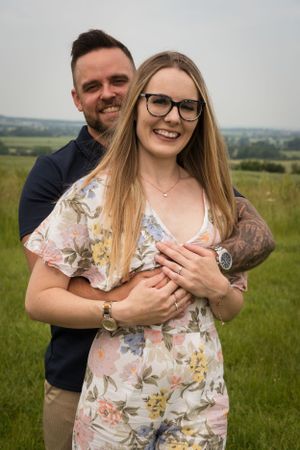 A newly engaged couple are having a cuddle. He is stood behind her with his arms wrapped around her. She is showing off her new ring.