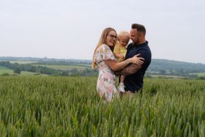 A mum and dad are holding their little girl, having a big cuddle in a large green field.