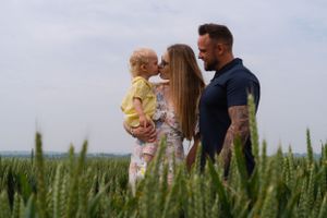 A mum is holding her little girl, giving her a kiss, whilst the dad is standing with his arm around the mum. In a large green wheat field.
