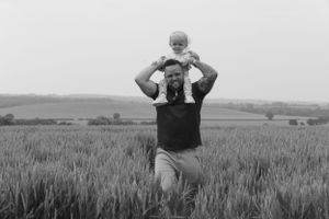 Black and white image of a dad and his little girl walking through a wheat field. The little girl is on the dads shoulders.