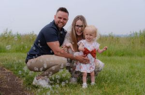 A mum, dad and little girl are crouched down playing with bubbles outside in a green field.