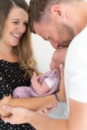 New parents holding their newborn baby girl between them. Dad kissing the baby on the head whilst mum is watching and smiling.
