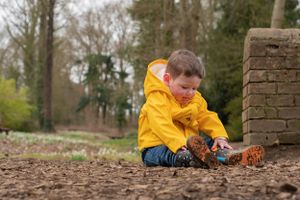 A little boy is sat in the leaves, with his yellow rain coat on and his wellies. He is looking down and playing with the leaves.