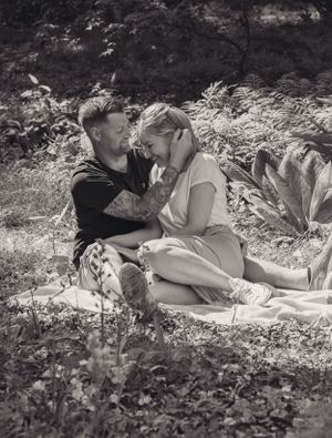A black and white image of a young couple sat down on the floor, in a garden. They are cuddling and he is tucking her hair behind her ear.