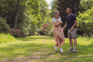 A young couple are dancing and laughing with each other, whilst looking at each other, in a beautiful green area.