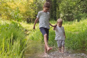 Two young boys playing in a stream through a green field, splashing with no shoes on. The brothers are holding hands.