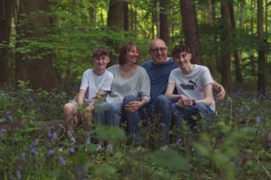 A young family are sat on a wooden log in a woods. They are all having a hug and are surrounded by bluebells.