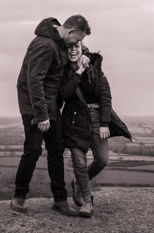 A married couple are having a dance and a cuddle on the hills with stunning views behind them. He is looking down at her and both laughing