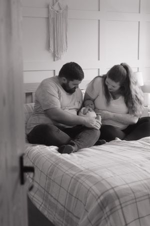 New parents are sat on their bed, holding their newborn baby. The photo is black and white.