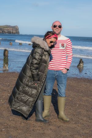 Couple walking along the beach, wearing winter coats and wellies and laughing.