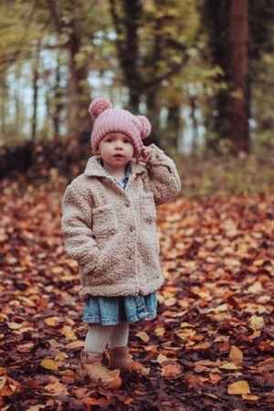 Little girl in autumnal leaves, pulling at her winter hat.
