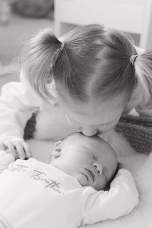 A black and white image of a little girl giving her baby brother a kiss on his head, whilst he sleeps.