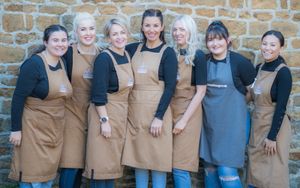 A group of beauticians stood in a line in front of a brick wall. They are all wearing brown aprons.