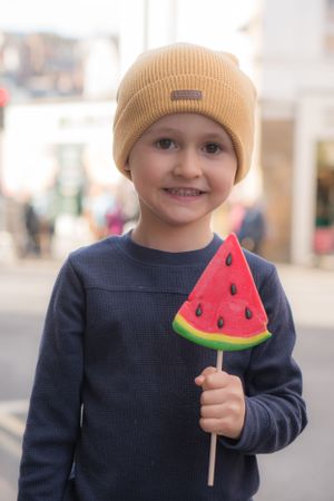 Little boy smiling at camera, holding a lolly in the style of a slice of watermelon.