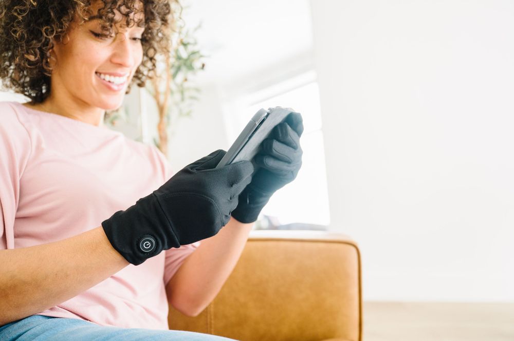 Person wearing a pink shirt and gloves using a mobile phone while sitting on a tan leather couch.