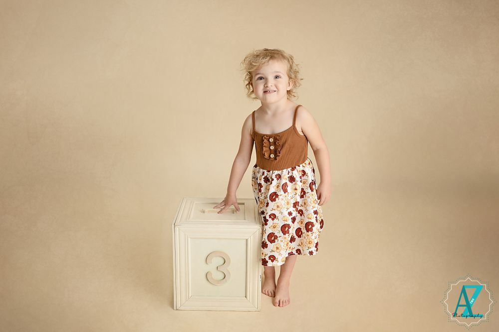 Toddler in brown top and floral skirt poses on white numbered cube against neutral background.